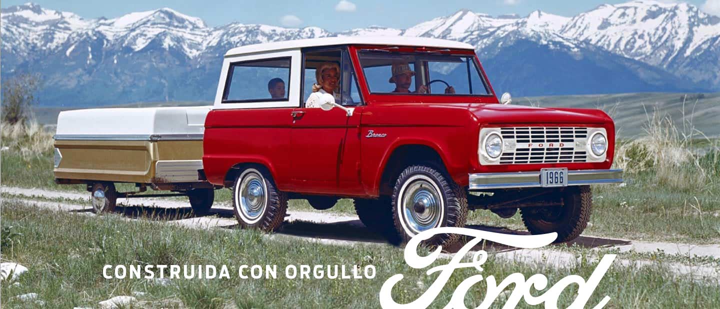 Una familia andando en un Ford Bronco Sport Wagon 1966 en Royal Maroon con techo Wimbledon White remolcando una casa rodante en un sendero doble por las montañas