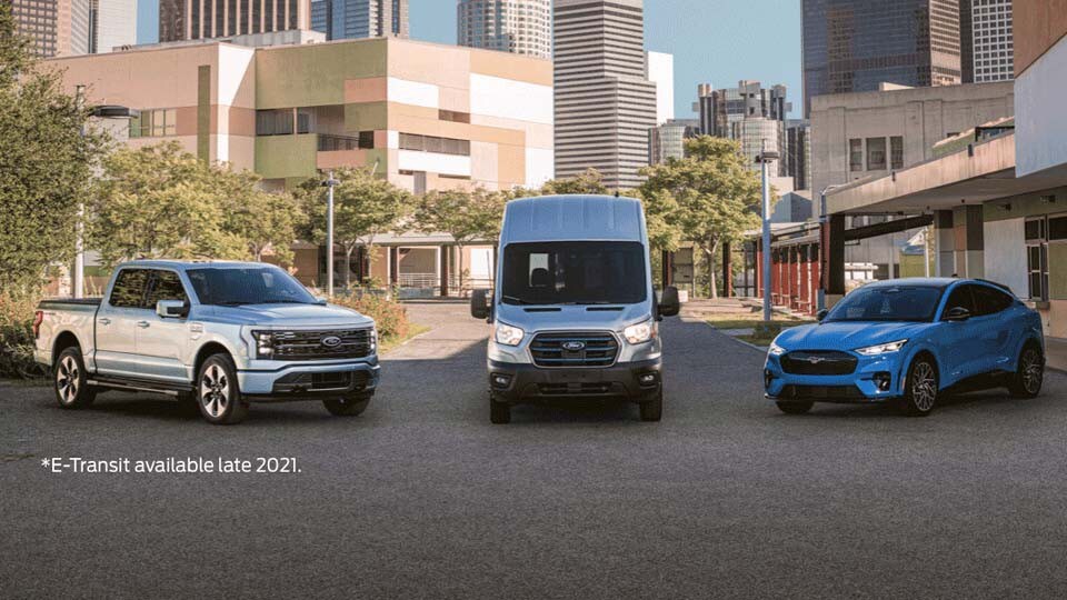 Una Ford Mustang Mach-E 2022, Ford F-150 Lightning 2022 y Ford E-Transit 2022 estacionadas en frente de una gran ciudad
