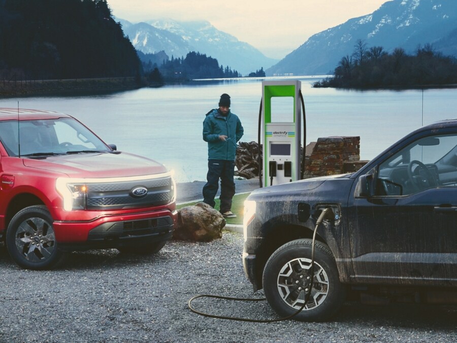 A la izquierda, una Ford F-150® Lightning® 2023 red. A la derecha, una black enchufada en un cargador público. Un lago a la distancia
