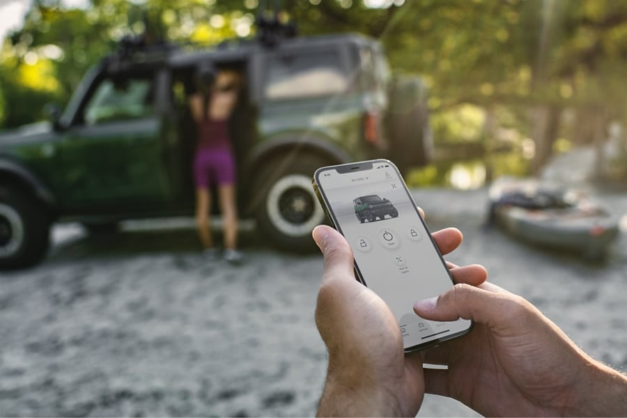Una persona sostiene un iPhone con la Aplicación FordPass en la pantalla y se ve una SUV Ford Bronco está estacionada de fondo.
