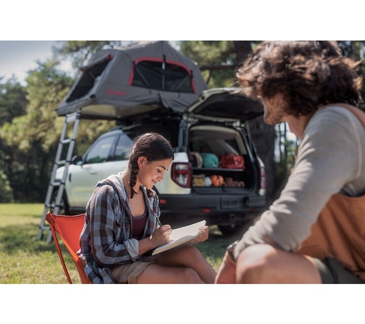 Una Ford Bronco® Sport 2024 de fondo, una mujer escribiendo en su diario y un hombre sentado cerca suyo
