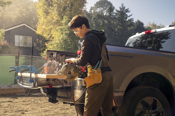 Un hombre cortando una tabla sujeta a la puerta trasera de una Ford F-150 Lightning® 2023