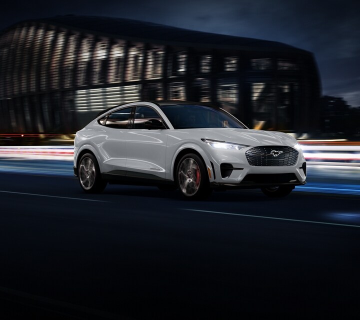 Una SUV Ford Mustang Mach-E 2023 en Space White Metallic circulando por una carretera frente a un estadio por la noche