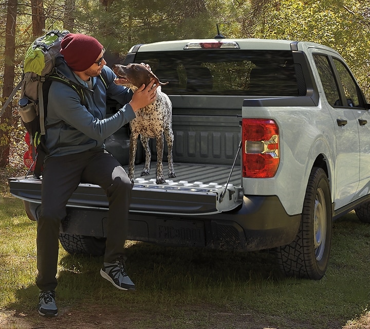 Una camioneta Ford Maverick® 2024 en Cactus Gray está estacionada en un lugar pintoresco con una persona y un perro en la plataforma