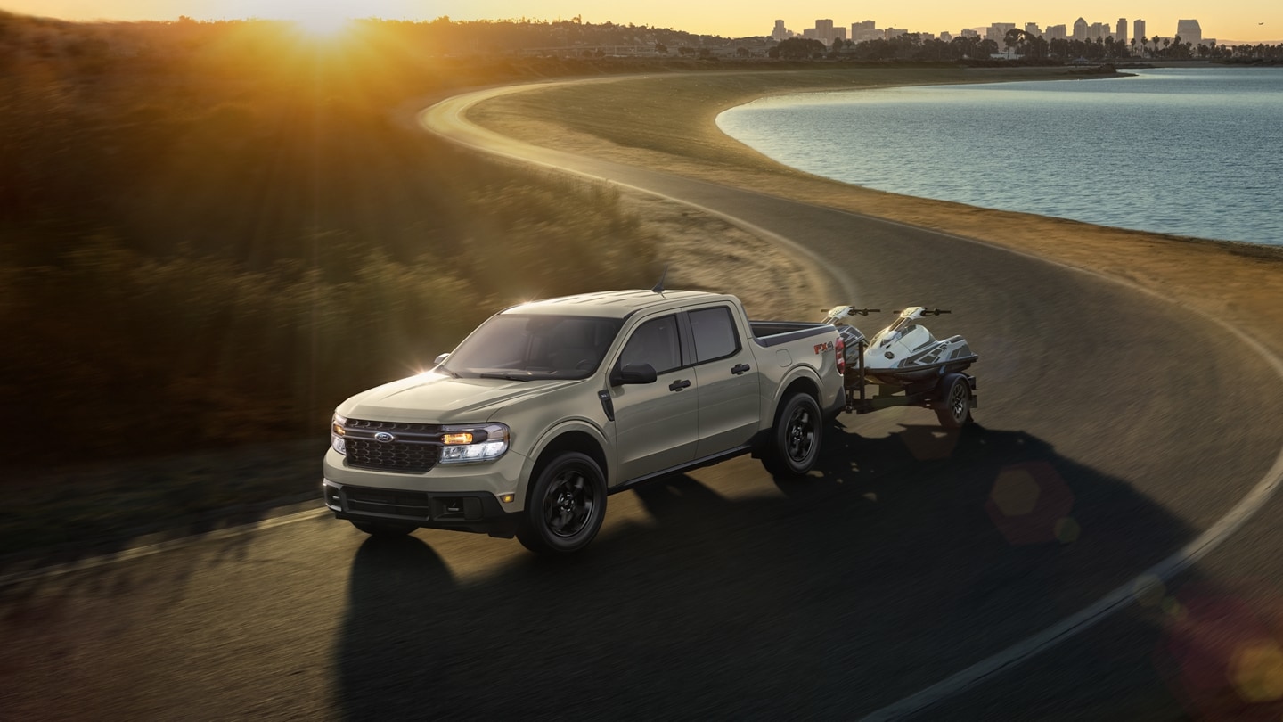Una Ford Maverick® 2024 remolcando jet skis en una carretera metropolitana al atardecer cerca del agua