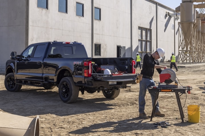 Hombre usando una sierra ingletadora en un lugar de trabajo con una camioneta Ford Super Duty® 2024 estacionada cerca con la puerta trasera baja